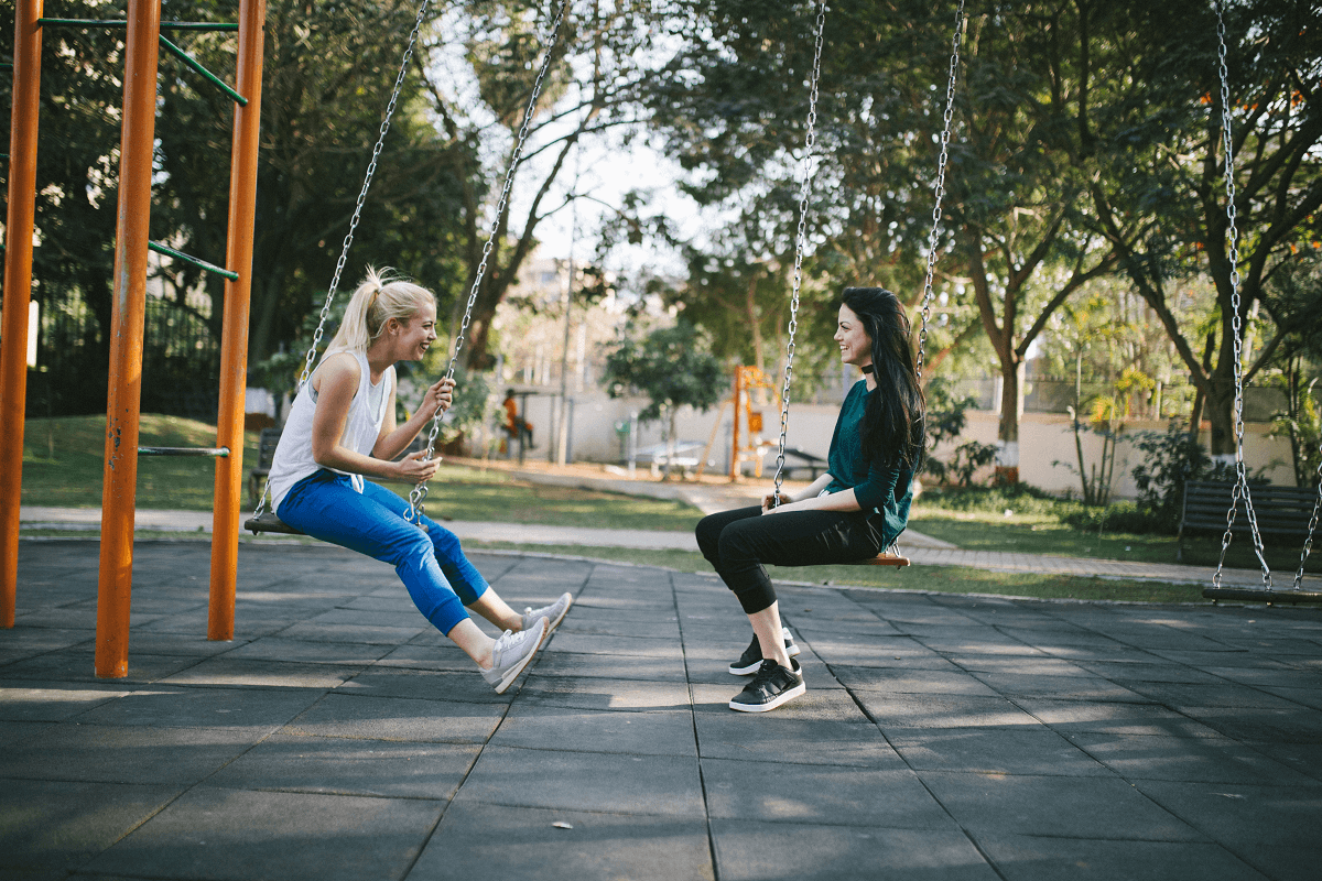 Two women on playground swings facing each other smiling